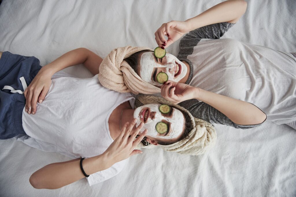 Two women relaxing on a bed with spa face masks and cucumbers on their eyes