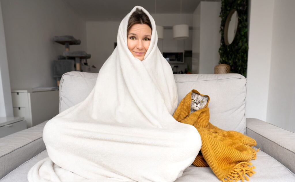 Woman cozy on a couch with blanket wrapped around her head and cat next to her with its own blanket wrapped around its head