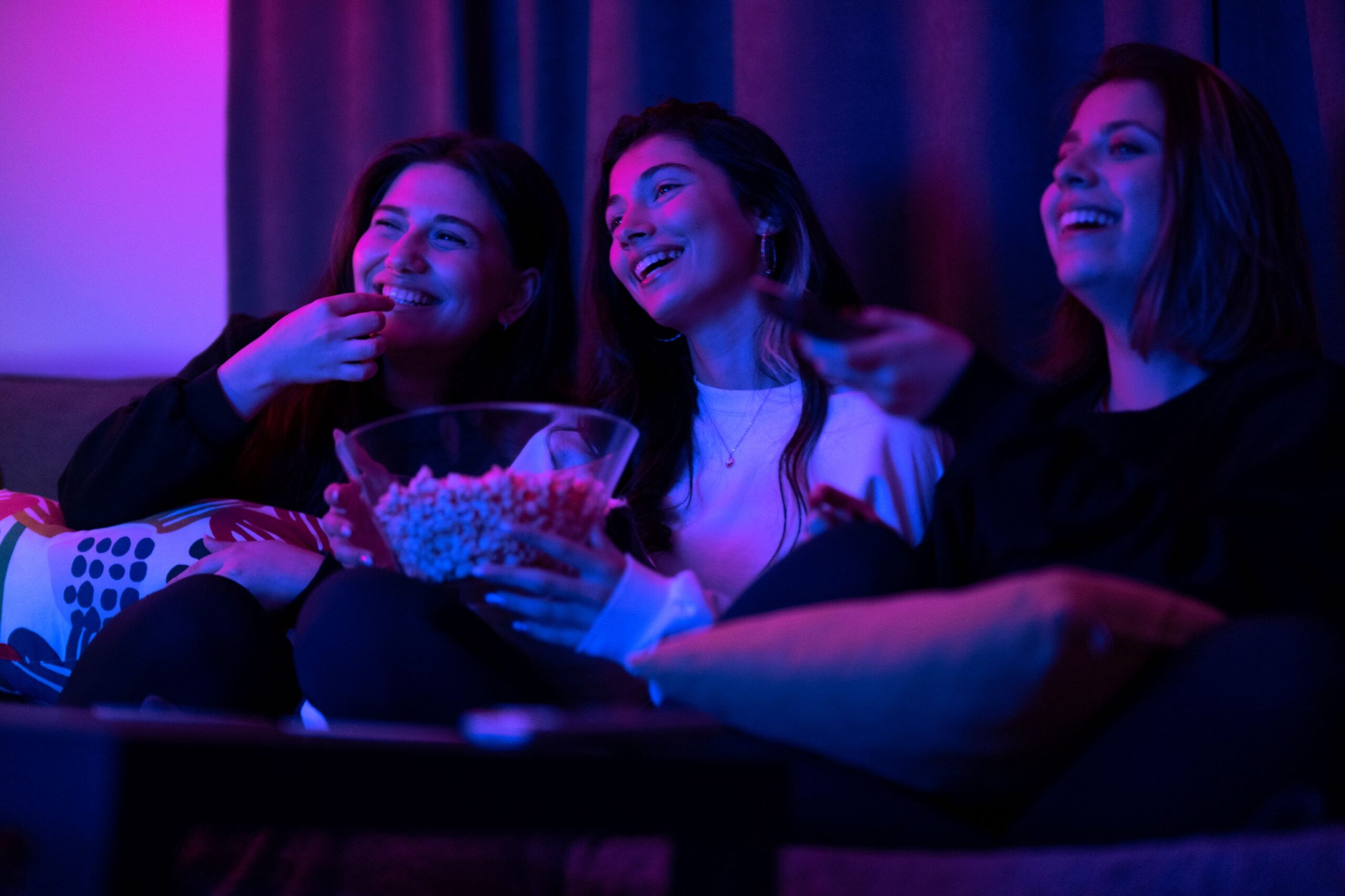 Three women sitting on a couch laughing and eating popcorn together in purple ambiance