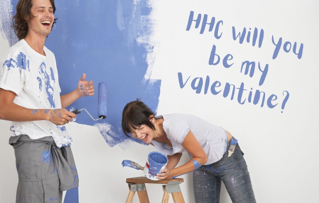 Couple laughing together while painting "HHC will you be my valentine?" on a white wall in blue paint