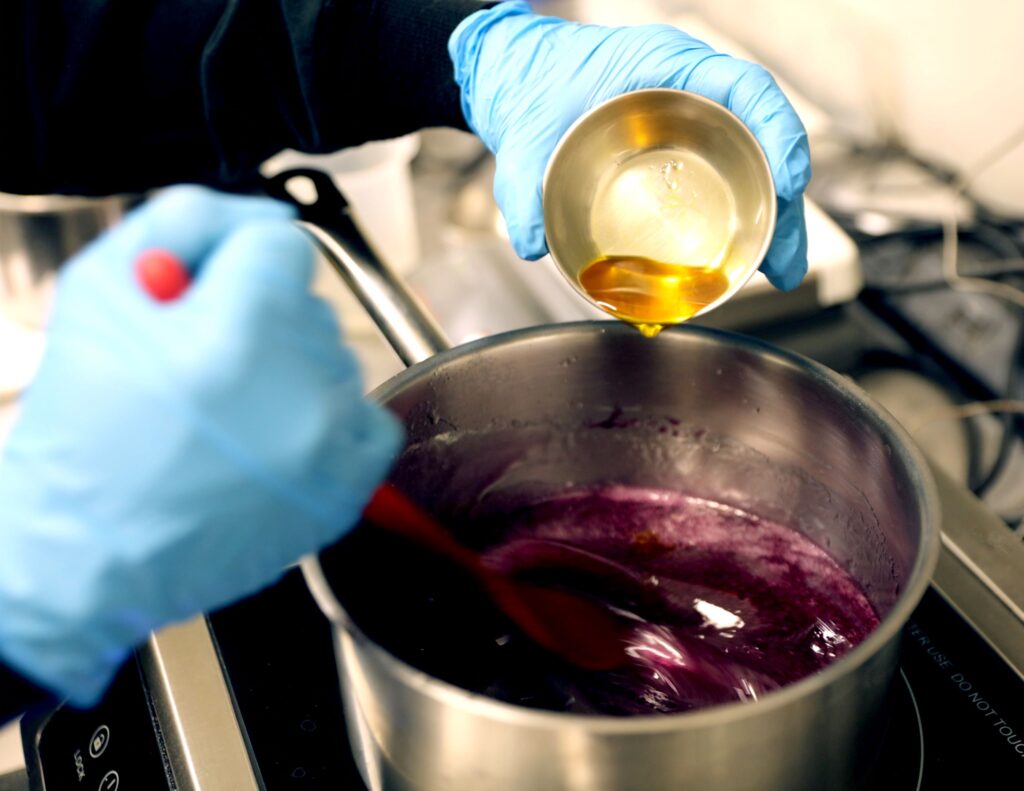 Gloved hands pouring mixture into large pot on a burner to create delta-9 gummies in a lab setting