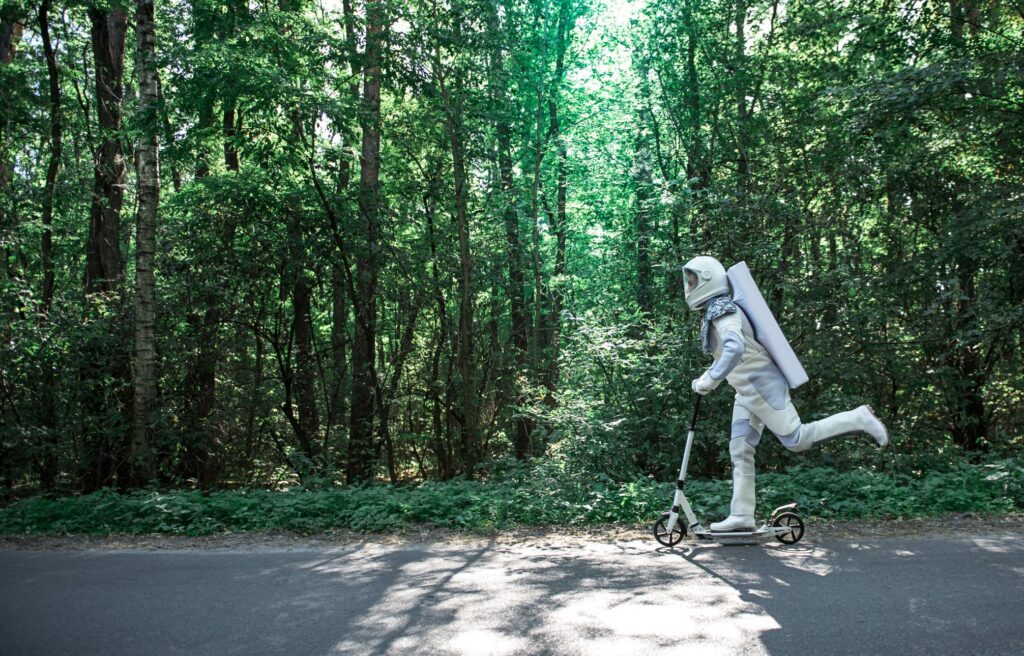Astronaut riding a scooter on a road in front of trees