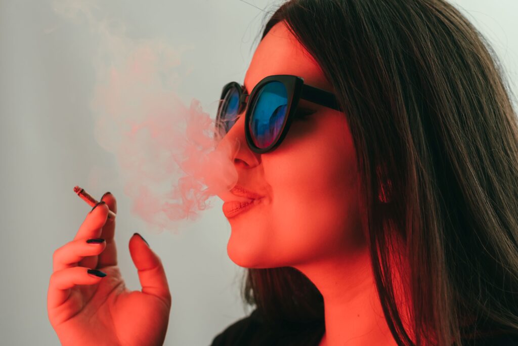 A woman wearing sunglasses and blowing out smoke from a pre-roll