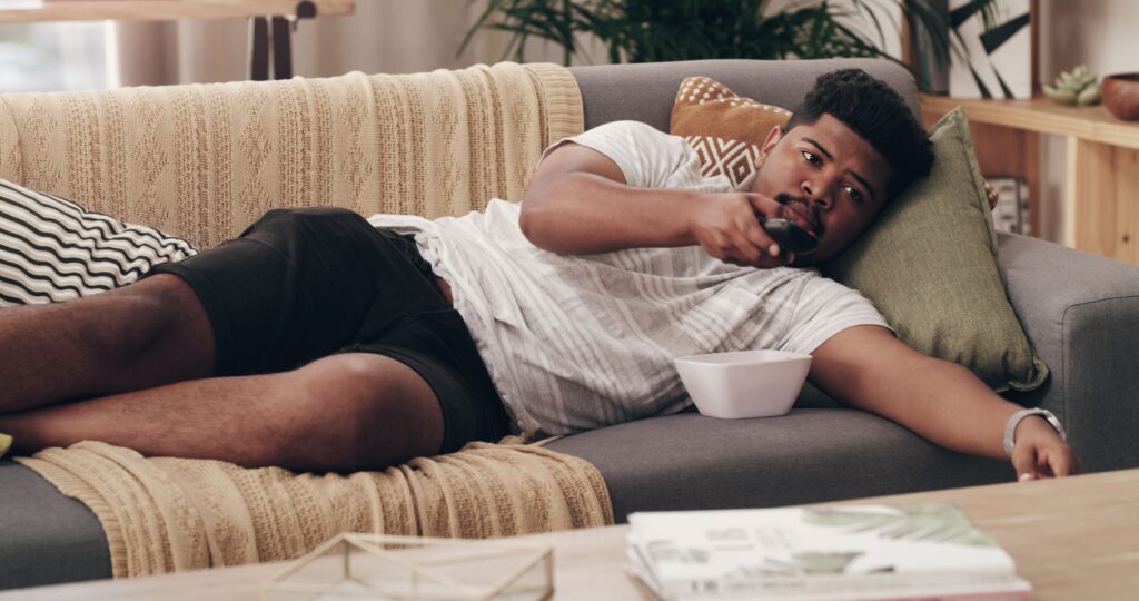 A man laying on the couch with a bowl of food and a TV remote in his hand